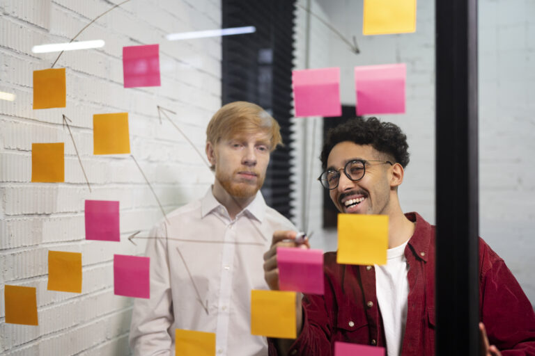 Duas pessoas sorrindo para um quadro de vidro com alguns post-its